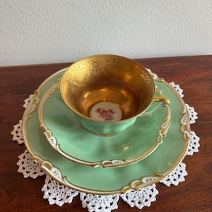 May include: A mint green porcelain tea cup and saucer set with a matching plate. The interior of the cup is gold-toned with a small floral motif at the base. The edges of the cup, saucer, and plate feature intricate white and gold filigree detailing.