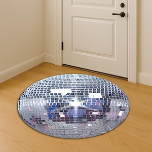 Op de afbeelding: Een ronde, zilverkleurige discoball-vloermat met een spiegelend oppervlak. De mat is gemaakt van kleine, vierkante spiegeltegels en ligt op een houten vloer voor een witte deur.