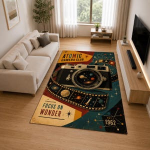 Puede incluir: Una alfombra rectangular con un gráfico de cámara vintage y una tira de película que rodea el objetivo. El diseño incluye el texto ATOMIC CAMERA CLUB, FOCUS ON WONDER y EST. 1962, con ilustraciones espaciales de planetas y estrellas.
