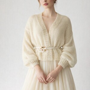 May include: Ivory-colored knit cardigan with a V-neck and long, voluminous sleeves. A white belt with a gold-toned buckle cinches the waist. The cardigan is paired with a flowing, cream-colored skirt, creating a soft, elegant look.