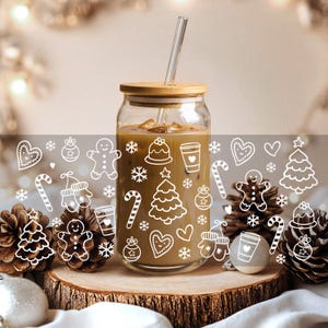May include: A clear glass tumbler with a bamboo lid and a glass straw, filled with a coffee drink. The glass is decorated with white Christmas-themed illustrations. The tumbler sits on a wooden slice, with pinecones and ornaments.