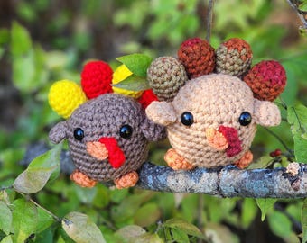 May include: Two adorable crochet turkey figurines perched on a branch. One is brown, the other is tan, both with black eyes, red wattles, and colorful tail feathers. The turkeys are surrounded by green foliage.