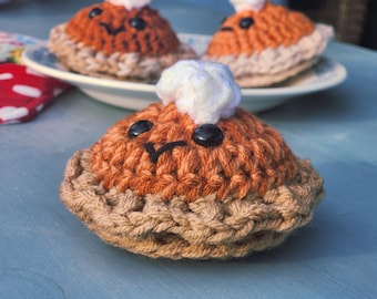 May include: Close-up of a crocheted pie with a smiling face and a dollop of white yarn "whipped cream." The pie has a brown crust and an orange filling. Other pies are visible in the background on a white plate.