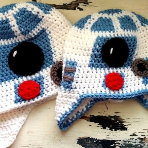 Könnte beinhalten: Zwei handgefertigte Häkelmützen im Stil eines bekannten Droiden-Charakters. Die Mützen sind weiß mit blauen und schwarzen Akzenten, mit einem großen schwarzen Auge, einem roten Kreis und grauen Details. Die Mützen sind so konzipiert, dass sie die Ohren bedecken.