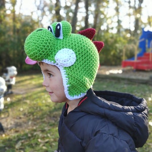 Può includere: Un bambino indossa un cappello Yoshi a uncinetto verde, bianco e rosa. Il cappello ha una grande testa di Yoshi verde con occhi neri e una lingua rosa.