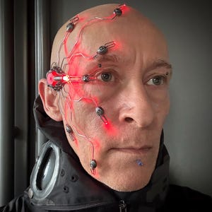 May include: A bald person wearing a black jacket with a futuristic cybernetic device attached to their face. The device features red glowing wires and small metallic components, creating a sci-fi aesthetic.