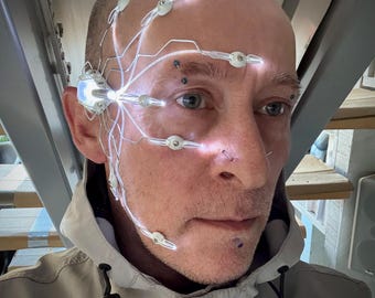 May include: A person wearing a futuristic headpiece with glowing lights and wires. The device is attached to the face and head, with small lights positioned around the eyes and forehead. The person is wearing a light-colored jacket.