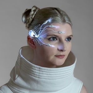 Puede incluir: Una mujer con un tocado futurista con luces blancas brillantes. El tocado tiene un diseño complejo con múltiples luces pequeñas que se extienden por la frente y alrededor de la oreja. Lleva una parte superior blanca de cuello alto.