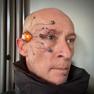 May include: A facial prosthetic device featuring a metallic, spider-like structure with thin, branching wires extending across the temple and forehead. It includes a central glowing orange light, small metallic spheres, and blue and red accents.