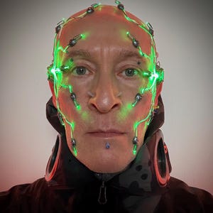 以下が含まれることがあります： 黒いジャケットを着た人物と未来的なヘッドピース。ヘッドピースは、顔と頭にワイヤーで接続された緑色の光を発しています。その人は頭を剃っており、唇にピアスをしています。