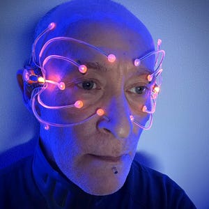 Könnte beinhalten: Eine Person, die ein futuristisches Headset mit leuchtenden orangefarbenen Kugeln und Drähten trägt. Das Headset ist an den Ohren und um die Augen befestigt, mit einem dunkelblauen Hintergrund. Die Person trägt ein dunkelblaues Oberteil.