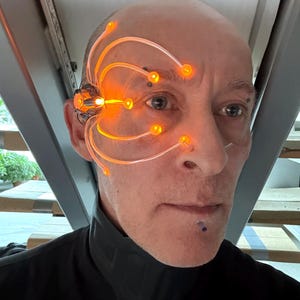 Könnte beinhalten: Nahaufnahme einer Person mit einem futuristischen, orangefarben leuchtenden kybernetischen Augengerät. Das Gerät hat mehrere leuchtende Ranken, die sich über das Gesicht erstrecken. Die Person trägt ein schwarzes Oberteil.