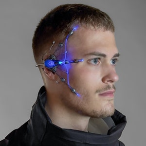 Puede incluir: Un dispositivo cibernético futurista con luces LED azules está adherido al lado de la cabeza de una persona. El dispositivo tiene un diseño mecánico complejo con múltiples puntos de fijación y acentos brillantes.