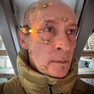 May include: A man wearing a gold puffer jacket with a futuristic headpiece. The headpiece has glowing orange lights and intricate wiring across the face and head. The background is a neutral industrial setting.