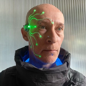 Pode incluir: Uma pessoa usando um capacete futurista com gavinhas e luzes verdes brilhantes. O capacete é preso ao lado da cabeça e emite uma luz verde brilhante. A pessoa está vestindo uma jaqueta preta com uma gola azul brilhante.