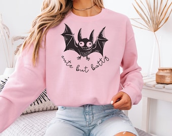 May include: A pale pink sweatshirt with a black cartoon bat design. The bat has large eyes and wings, with the words "cute but batty" written below. The sweatshirt has a crew neck and long sleeves.