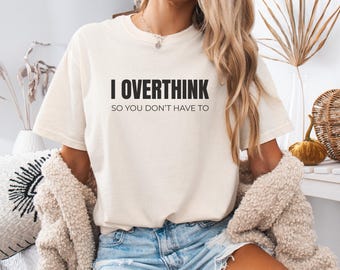 Pode incluir: Camiseta creme com o texto preto "I OVERTHINK SO YOU DON'T HAVE TO". A camiseta é combinada com calções de ganga e um cardigan bege. O visual geral é casual e confortável.