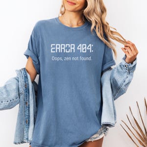 May include: A blue t-shirt with the text "ERROR 404: Oops, zen not found" in white. The shirt has short sleeves and a relaxed fit. A denim jacket is draped over the shoulders.