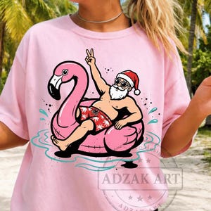 May include: A light pink t-shirt featuring a graphic of Santa Claus wearing sunglasses and red patterned swim trunks, sitting on a large pink flamingo pool float. Santa is making a peace sign with one hand. The text ADZAK ART is visible.
