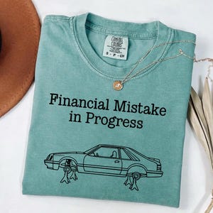 Könnte beinhalten: Ein teal-farbenes Comfort Colors T-Shirt mit dem schwarzen Aufdruck "Financial Mistake in Progress" und einer Strichzeichnung eines Autos auf Wagenhebern. Eine silberne Halskette liegt auf dem Shirt.
