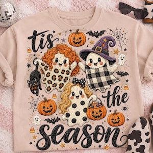 May include: A beige crewneck sweatshirt featuring a graphic print with the text 'tis the season'. The design includes three cartoon ghosts, one with leopard print, one with buffalo plaid and a purple hat, and one with polka dots, plus pumpkins, a black cat, bats, and spiderwebs.