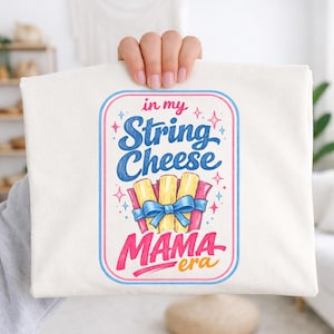 May include: A white t-shirt featuring a colorful graphic. The design includes the text "in my String Cheese Mama era" in blue, pink, and yellow, with illustrations of string cheese and a blue bow.