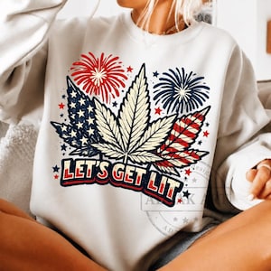 Op de afbeelding: Een wit sweatshirt met ronde hals en een grafische print van drie cannabisbladeren. De bladeren zijn versierd met rood-wit-blauwe Amerikaanse vlagpatronen en vuurwerk. De tekst "LET'S GET LIT" is gedrukt in dikke, verweerde rode en blauwe letters.