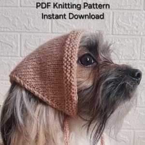 May include: A dog wearing a knitted, light brown triangular hat with matching ties. The text "PDF Knitting Pattern Instant Download" is visible above the dog. The dog has long, multi-coloured fur.