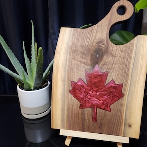 May include: Wooden chopping board with a handle and a red maple leaf design. The maple leaf is inlaid with a glossy, swirled red material. The board is displayed on a small wooden easel.