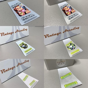 May include: White and silver stickers featuring classic arcade game graphics. One sticker displays "AMIDAR" with a cartoon character, while others show "FROGGER" and "FROGGER II THREE DEEP" logos. The stickers are next to a box with "Vintage Arcading" text.