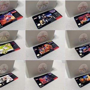 May include: Nine retro video game cartridges with colorful game art and text, including titles like "Batman," "Double Dragon," and "Darkwing Duck." The cartridges are black with red accents and are displayed near a white box with a logo.