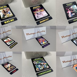 May include: A collection of vintage arcade game cartridges, each with a unique game title like "Black Jack," "Breakout," and "Casino." The cartridges are black with colorful game art and text, displayed with matching title labels and white boxes.