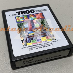 May include: Black Atari 7800 video game cartridge featuring the game Klax. The cartridge has a white label with colorful graphics and the Atari 7800 logo. The game title and Atari logo are visible.