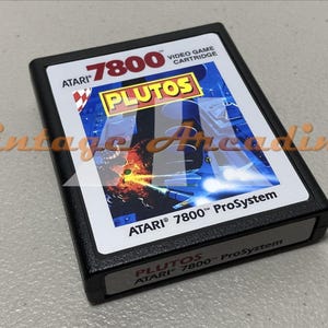 May include: Black Atari 7800 ProSystem video game cartridge for the game "Plutos." The cartridge has a white label with the game title and Atari branding. The game's cover art features a space battle scene with a blue and orange color scheme.