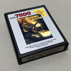 May include: A black Atari 7800 Super Game Cartridge for the ProSystem, featuring the game "Missing in Action." The cartridge has a white label with game art and text. Copyright 1989, TNT Games. Printed in Hong Kong.
