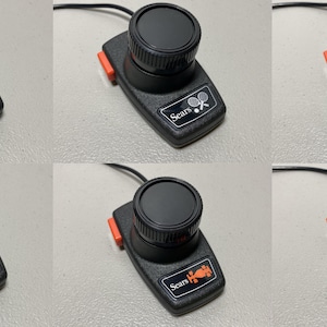 May include: Black Atari and Sears video game controllers with a black knob, orange accents, and a black cable. The controllers have various game icons and text, including "Atari", "Sears", "paddle", and "driving".