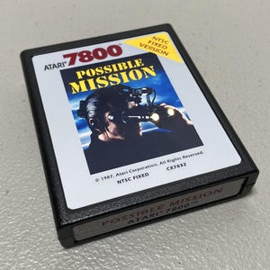 May include: A black Atari 7800 game cartridge for "Possible Mission." The label features a man wearing a headset and holding a device, with the game title in yellow and blue. The cartridge has the text "NTSC FIXED VERSION."