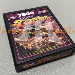 May include: A black Atari 7800 Super Cobra video game cartridge. The label features the game title in yellow and orange, with a background of a fiery sky and aircraft. Text on the label reads "High-flying 3-D combat action! You're in control!"