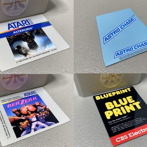 May include: Four vintage Atari 5200 game cartridge labels. Titles include "Asteroids", "Berzerk", "Astro Chase", and "Blueprint". The labels feature colorful graphics and text, with the Atari logo prominently displayed.
