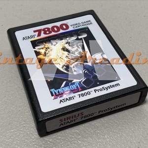 May include: Black Atari 7800 video game cartridge for the game Sirius. The cartridge has a white label with the game title and a graphic of a spaceship and explosions. The Atari logo is visible.