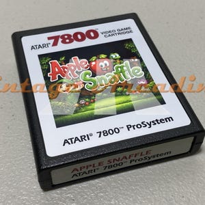 May include: A black Atari 7800 video game cartridge featuring the game "Apple Snaffle." The cartridge has a white label with the game title and colorful graphics of cartoon characters. The Atari logo is visible.