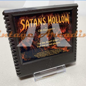 May include: A black Atari 5200 game cartridge for "Satan's Hollow" by CBS Electronics. The label features fiery orange and red graphics with the game title in bold, stylized lettering. The cartridge is displayed on a clear stand.