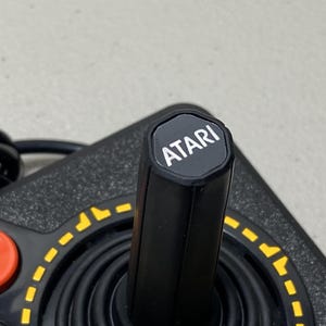 May include: Close-up of a black Atari joystick controller. The joystick has a black shaft with the Atari logo on top. A red button and yellow dashed lines are visible on the base.
