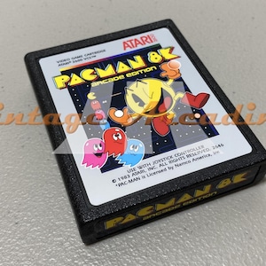 May include: A black Atari Pac-Man 8K Arcade Edition video game cartridge. The label features Pac-Man and ghosts in a colorful design. The text "PAC-MAN 8K arcade edition" is visible.