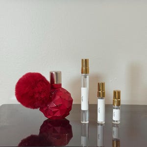 Peut inclure: Un flacon de parfum rouge avec un design facetté et un pompon rouge duveteux, à côté de trois flacons d'échantillons de parfum transparents avec des bouchons dorés. Les flacons ont des étiquettes blanches.
