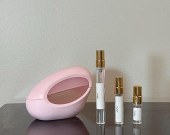 May include: A pink, oval-shaped perfume case and three perfume vials with gold caps. The case has a large, oval opening. The vials are clear with white labels and gold caps. The perfume bottles are different sizes.