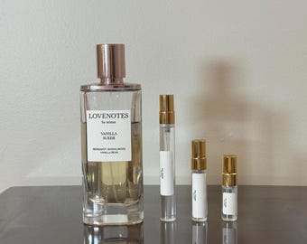 以下が含まれることがあります： ローズゴールドのキャップが付いた透明な香水瓶と、「LOVENOTES by ariana VANILLA SUEDE BERGAMOT SANDALWOOD VANILLA BEAN」と書かれた白いラベル。 金色のキャップと白いラベルが付いた3つの小さな香水瓶も写っています。