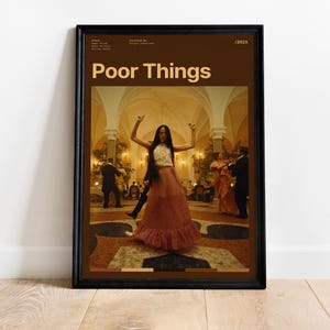 May include: Framed poster featuring the movie title "Poor Things" in a brown frame. The poster depicts a woman in a long pink dress dancing in a ballroom. Other people are in the background. The poster is displayed against a white wall.