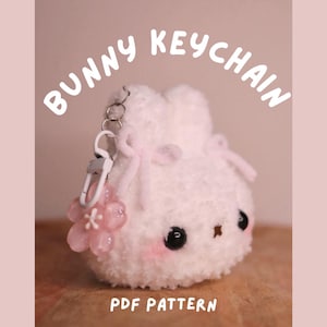 Può includere: Un portachiavi a forma di coniglietto bianco e soffice con un ciondolo a forma di fiore rosa. Il portachiavi ha occhi neri, guance rosa e un piccolo naso marrone. Il testo "BUNNY KEYCHAIN" è sopra il coniglietto, e "PDF PATTERN" è sotto.