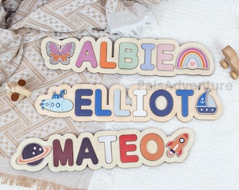 May include: Wooden name puzzles spelling "ALBIE", "ELLIOT", and "MATEO". Each puzzle features colorful letters and themed illustrations, including a butterfly, rainbow, submarine, sailboat, planet, and rocket. The puzzles are on a neutral-toned fabric.